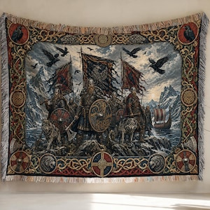 Op de afbeelding: Een geweven wandtapijtdeken met een gedetailleerde scène van krijgers met schilden, bijlen en vaandels op een rotsachtige kust met wolven en raven. De rand toont ingewikkelde knoopwerkpatronen, ronde schilden en diermotieven met franjes.
