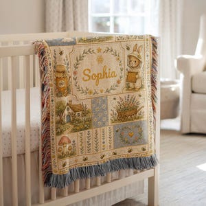 May include: A woven throw blanket featuring a patchwork design with illustrations of a rabbit, a honey pot, a cottage, a wheelbarrow with flowers, and mushrooms. The name Sophia is centered in a floral wreath. The blanket has blue fringe.