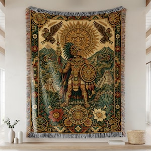 Op de afbeelding: Een geweven wandkleed met een centrale Azteekse krijgersfiguur die een speer en schild vasthoudt, omringd door een grote zon, adelaars, slangen en piramides. Het ontwerp bevat bloemmotieven, geometrische randen en een franje aan de onderkant.