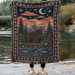 Puede incluir: Una manta tejida que muestra un cielo nocturno azul oscuro con una luna creciente y estrellas sobre un paisaje montañoso. Un oso pardo se encuentra en un bosque cerca de un río con dos peces y un pato. El borde tiene un patrón repetitivo de piñas.