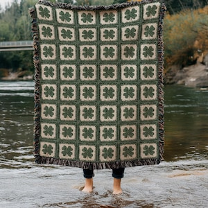Könnte beinhalten: Eine grün-beige gehäkelte Decke mit einem sich wiederholenden Muster aus vierblättrigen Kleeblättern. Die Decke hat einen Fransenrand und wird vor einer Person gehalten, die in einem Fluss steht.