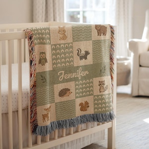 May include: A woven throw blanket featuring a patchwork design of sage green and beige squares. The blanket displays illustrations of a rabbit, bear, owl, skunk, hedgehog, raccoon, squirrel, and deer, with the name Jennifer embroidered in the center.