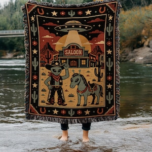 May include: A woven throw blanket featuring a desert scene with a UFO beaming light onto a building labeled SALOON. The design includes a gray alien wearing a cowboy hat and bandana standing next to a gray horse, surrounded by cacti and stars.