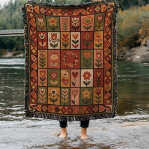 Puede incluir: Una manta tejida que presenta una cuadrícula de paneles rectangulares con motivos florales, de corazones y de aves en tonos rojos, verdes, amarillos y naranjas. La manta tiene un borde geométrico decorativo y flecos en los cuatro lados.