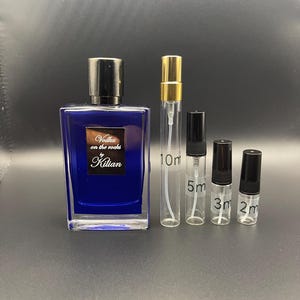 Könnte beinhalten: Ein rechteckiger, dunkelblauer Parfümflakon mit schwarzem Verschluss, mit dem Text "Vodka on the rocks" und "Kilian" in Weiß. Daneben vier transparente Glasfläschchen mit den Aufschriften 10ml, 5ml, 3ml und 2ml, mit goldenen und schwarzen Verschlüssen.