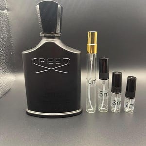 Op de afbeelding: Een zwarte parfumfles met het woord "CREED" in witte letters en verschillende kleine glazen flesjes met zwarte en gouden doppen. De flesjes zijn gelabeld met metingen van 2 ml, 3 ml, 5 ml en 10 ml.