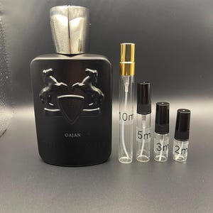 May include: A black perfume bottle with a silver cap and the word "Oajan" printed on the front. The bottle is flanked by four clear glass vials with black caps, labeled 10m, 5m, 3m, and 2m. The perfume bottle has a crest with two horses.