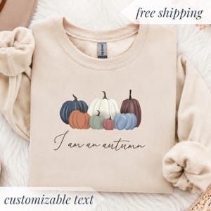 May include: A beige crewneck sweatshirt featuring a graphic of six pumpkins in various colors including blue, orange, white, green, and dark red. Below the pumpkins, the text reads I am an autumn in a dark brown cursive script font.