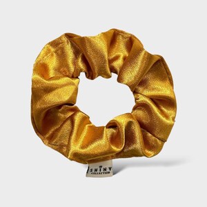 Könnte beinhalten: Ein goldgelbes Haargummi aus glänzendem Stoff. Das Haargummi hat eine kreisförmige Form mit einem gerafften Design. Ein kleines weißes Etikett mit dem Text "SHINY COLLECTION" ist angebracht.