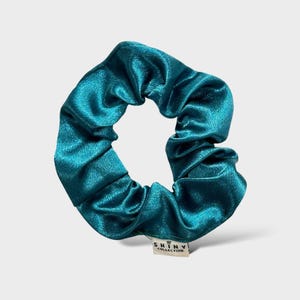 Könnte beinhalten: Türkisfarbene Satin-Haargummi. Das Haaraccessoire besteht aus einem glänzenden, gerafften Stoff. Ein kleines weißes Etikett mit dem Text "SHINY COLLECTION" ist am Haargummi befestigt.