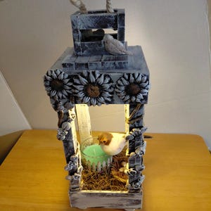 May include: A decorative wooden lantern with a rope handle, painted in distressed white and gray. The lantern features sunflower accents, a small bird figurine, and a candle holder with a green candle inside. The lantern is illuminated.