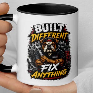 May include: A white ceramic mug with a black handle and rim, featuring a graphic of a bulldog wearing a cap and holding a wrench. The text reads BUILT DIFFERENT and FIX ANYTHING, surrounded by mechanical gears and a red toolbox illustration.