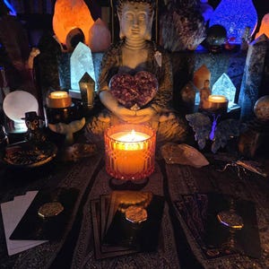 May include: A serene scene featuring a seated Buddha statue holding a heart-shaped amethyst. A lit candle in a decorative glass holder sits in front of the statue. Tarot cards and crystals are arranged around the candle, creating a spiritual ambiance.