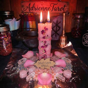 May include: A lit pink candle with dried rose petals and a pink string wrap. The candle is surrounded by rose petals, crystals, and a sign that reads "SELF-LOVE SPELL". The background includes jars, a bell, and a wooden sign that reads "Adrienne Tarot".