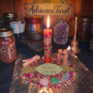 Op de afbeelding: Een brandende rode kaars met gouden accenten en gedroogde rozenblaadjes, omringd door een liefdesspreukarrangement. Het arrangement bevat een roze hartvormig kristal, een groen hartvormig kristal en een blad met de tekst "Avoidant Love Spell".