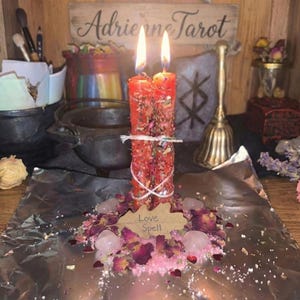 Op de afbeelding: Twee brandende rode kaarsen, versierd met gedroogde kruiden en vastgebonden met touw. Ze zijn omringd door rozenblaadjes, kristallen en een houten label met de tekst "Love Spell". Een bord erboven luidt "Adrienne Tarot".