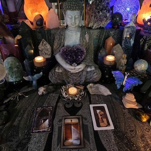 May include: A serene scene featuring a seated Buddha statue holding a heart-shaped amethyst. The setting includes crystals, candles, and tarot cards, creating a spiritual and decorative display. Various crystals and a blue butterfly sculpture are also present.