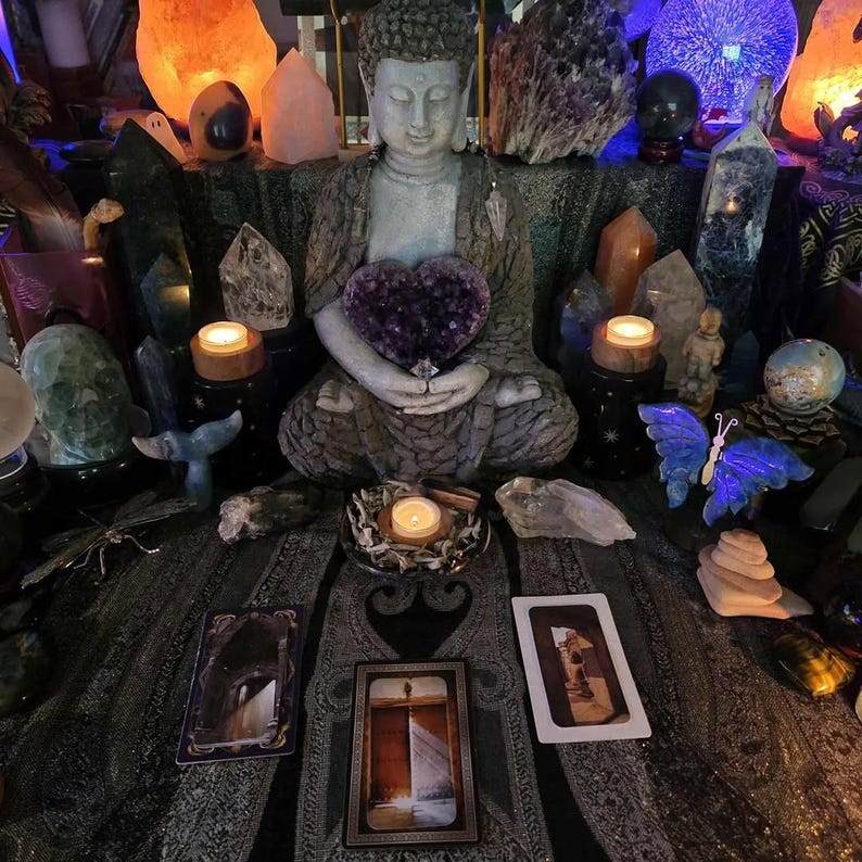 May include: A serene scene featuring a seated Buddha statue holding a heart-shaped amethyst. The setting includes crystals, candles, and tarot cards, creating a spiritual and decorative display. Various crystals and a blue butterfly sculpture are also present.