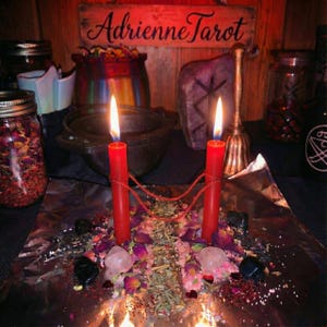 Può includere: Due candele rosse collegate da uno spago rosso su carta stagnola, circondate da petali di fiori secchi, erbe e piccoli cristalli. Un cartello in legno con il testo AdrienneTarot è sullo sfondo, insieme a barattoli e una campana di metallo.