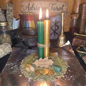 May include: Two lit taper candles, one green and one gold, are tied together with twine. They stand on a bed of dried herbs, gold-colored coins, bay leaves, and two crystals. A brown paper tag reads Road Opener & Financial Luck.