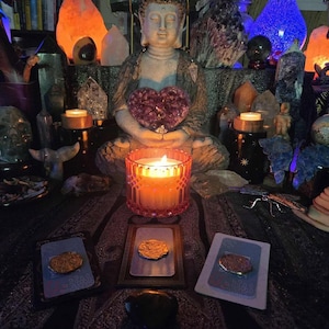 May include: A candle in a pink glass holder is lit, with a Buddha statue in the background holding a purple heart-shaped crystal. Tarot cards and crystals are arranged on a patterned cloth, with other candles and crystals in the background.