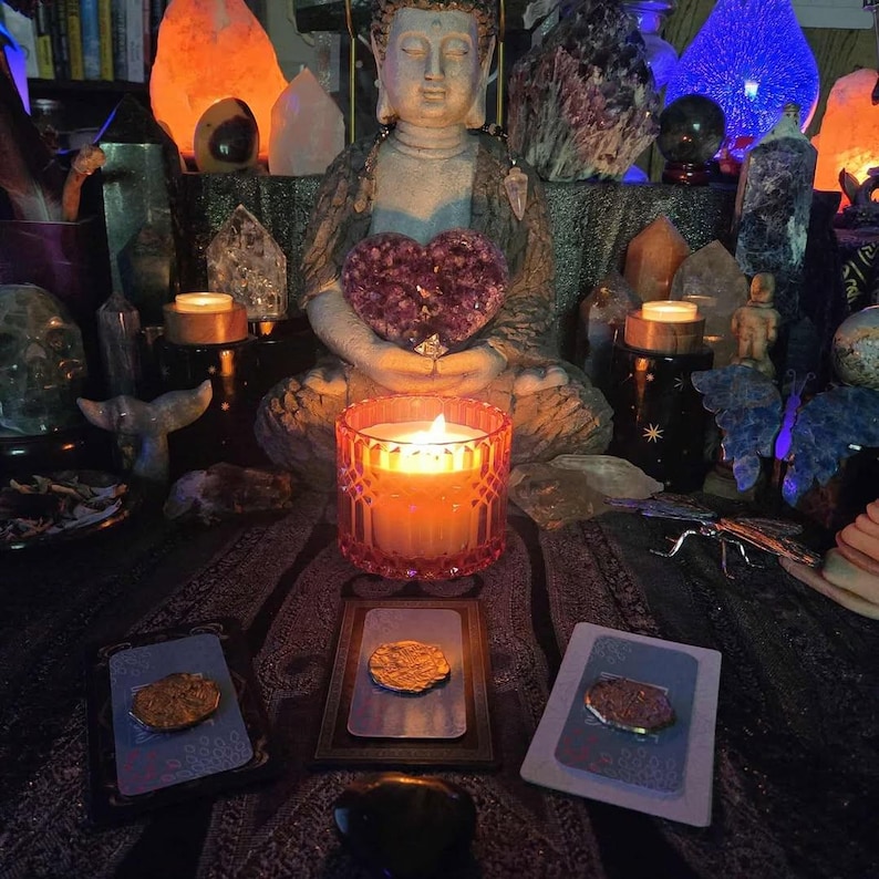 May include: A candle in a pink glass holder is lit, with a Buddha statue in the background holding a purple heart-shaped crystal. Tarot cards and crystals are arranged on a patterned cloth, with other candles and crystals in the background.