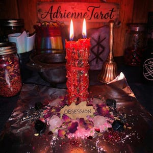 Può includere: Una candela rossa accesa, adornata con fiori ed erbe essiccate, su una superficie di alluminio. La parola "OBSESSION" è su un'etichetta di legno. Quarzo rosa e pietre nere circondano la candela. Un cartello di legno recita "Adrienne Tarot".