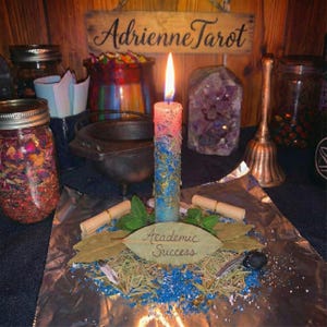 May include: A lit candle with blue, pink, and gold accents sits on a foil-covered surface, surrounded by herbs, scrolls, and a leaf that reads "Academic Success." The background includes jars, a cauldron, and a wooden sign that reads "Adrienne Tarot."