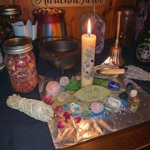 May include: A lit candle with purple accents and a leaf that says "Text Me Now!" sits on a table with crystals, herbs, and a small cauldron. Jars filled with dried flowers and herbs are also visible.