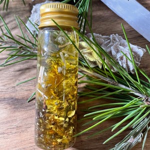 Könnte beinhalten: Eine kleine Glasflasche mit goldenem Deckel, gefüllt mit einer gelben Flüssigkeit und Kräutern. Die Flasche ist von grünen Kiefernnadeln, einem weißen Kristall und einer kleinen Felsformation umgeben. Die Gegenstände sind auf einer Holzoberfläche angeordnet.