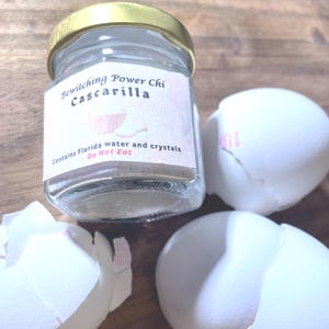 May include: A small glass jar with a gold lid, labelled "Bewitching Power Chi Cascarilla," is positioned near three cracked white eggshells. The label states the contents include Florida water and crystals, with a warning: "Do Not Eat."