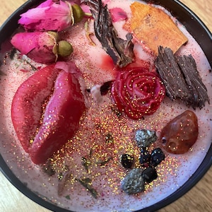 Könnte beinhalten: Eine dekorative Kerze in einem schwarzen Behälter mit einer rosa Wachsbasis. Die Kerze ist mit getrockneten Blumen, Glitzer und Edelsteinen verziert. Die Anordnung umfasst rosa Rosenknospen, eine rote Rose und verschiedene Kristalle.