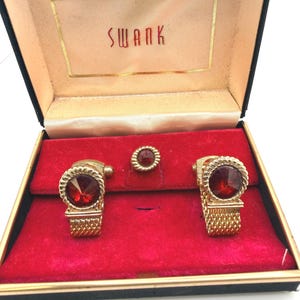 May include: Vintage gold-tone cufflink and stud set with a red gemstone center, presented in a black box with the word "SWANK" in gold lettering. The set includes two cufflinks and one stud, all with a matching design.