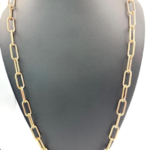 May include: A gold-colored chain necklace with elongated rectangular links. The necklace is displayed on a black mannequin, showcasing its design and length. The chain is a simple yet elegant piece of jewelry.