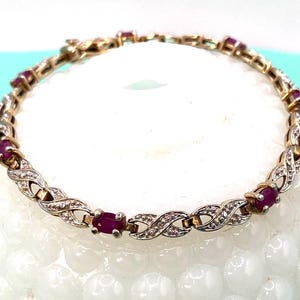 May include: A gold-toned bracelet featuring oval-shaped red gemstones and small, clear accent stones. The bracelet has an intricate design with a repeating pattern of intertwined links. The gemstones are evenly spaced along the bracelet's length.