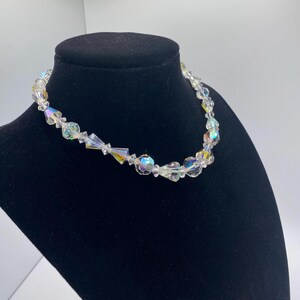 May include: A clear, iridescent beaded necklace with various shaped beads. The necklace is displayed on a black mannequin. The beads reflect light, creating a rainbow effect. The necklace is a choker style.