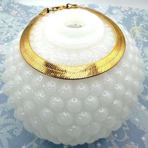 May include: A gold-colored herringbone chain bracelet is draped over a white, textured, round object. The bracelet has a classic design with a secure clasp. The background is a light blue and white patterned surface.