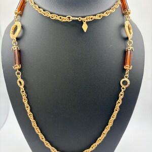 May include: A long gold-tone chain necklace featuring a series of cylindrical amber-colored resin beads and circular gold-tone metal links. The necklace includes a small diamond-shaped charm hanging from the center of the chain near the clasp.