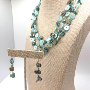 May include: A multi-strand necklace and matching drop earrings set featuring iridescent light blue and teal shell beads. The necklace consists of several strands of circular and square-shaped shell pieces interspersed with small blue glass beads.