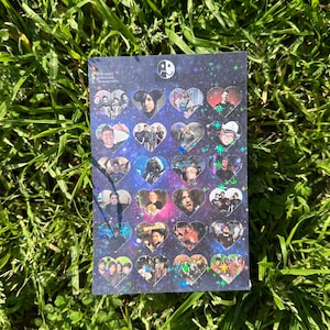 Op de afbeelding: Een rechthoekige stickervel met hartvormige afbeeldingen van mensen tegen een sterrenachtergrond. De stickervel heeft een holografische afwerking en een yin-yang symbool bovenaan. De vel ligt op groen gras.