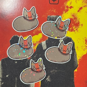 May include: Five cat stickers with a brown body, a red stripe across the forehead, and a white outline. The stickers have a holographic effect with colorful star-shaped sparkles. The background is red and yellow.