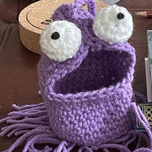 May include: A handmade, purple crochet monster with large white eyes and black pupils. The monster has an open mouth and fringe at the bottom. The crochet project is a whimsical, unique design.