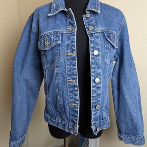 May include: A medium-wash denim jacket with a classic collar and button-up front. The jacket features two chest pockets and buttoned cuffs. The garment is displayed on a black mannequin, showcasing its structure and design. The jacket's color is a faded blue.
