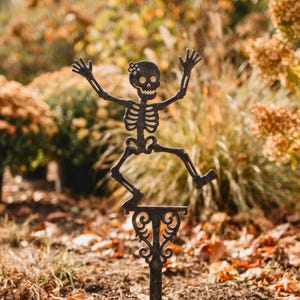 May include: A dark metal garden stake featuring a skeleton figure with arms raised and one leg bent. The skeleton is attached to a decorative scrollwork post that extends into a pointed base, with a small green pumpkin resting at the bottom.