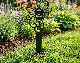 Könnte beinhalten: Ein schwarzer Metall-Gartenstecker mit einem stilisierten Bienen-Design. Die Biene hat detaillierte Flügel und einen Körper mit dekorativen Wirbeln. Der Stecker befindet sich in einem Garten mit grünem Gras und verschiedenen Blumen im Hintergrund.