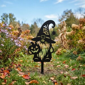 Peut inclure: Une décoration de jardin en métal noir en forme de gnome portant un chapeau haut avec une découpe en toile d'araignée et une araignée suspendue. Le gnome tient une citrouille sculptée avec un visage de lanterne. Design ajouré.