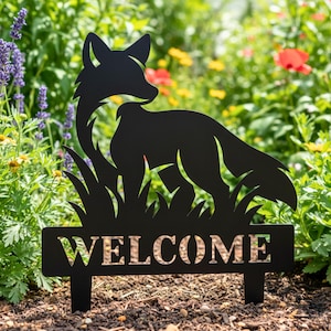 May include: Black metal garden stake featuring a fox silhouette standing in tall grass. The stake includes the word "WELCOME" cut out in the sign. The fox is facing to the right, with a detailed outline. The background shows a garden with colorful flowers.