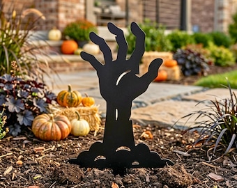 Könnte beinhalten: Schwarze Halloween-Dekoration aus Metall in Form einer aus dem Boden ragenden Zombiehand. Die Szene umfasst Kürbisse und Herbstlaub, was eine gruselige Szenerie erzeugt.