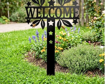 Könnte beinhalten: Schwarzes Metall-Gartenschild mit dem ausgeschnittenen Wort "WELCOME". Das Schild hat Blatt- und Sterndesigns mit einem vertikalen Pfosten zur Bodenaufstellung. Das Schild befindet sich in einem Garten mit grünem Gras und bunten Blumen.