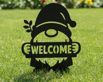 Könnte beinhalten: Schwarzes Metall-Gartenzwerg-Schild mit dem ausgeschnittenen Wort "WELCOME". Der Zwerg hat eine Zipfelmütze, einen Bart und einen Zweig mit Blättern. Das Schild steht im grünen Gras.